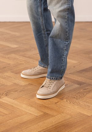 Gambe con jeans blu e scarpe da ginnastica beige in suede che stanno su un pavimento in legno con motivo all'interno.