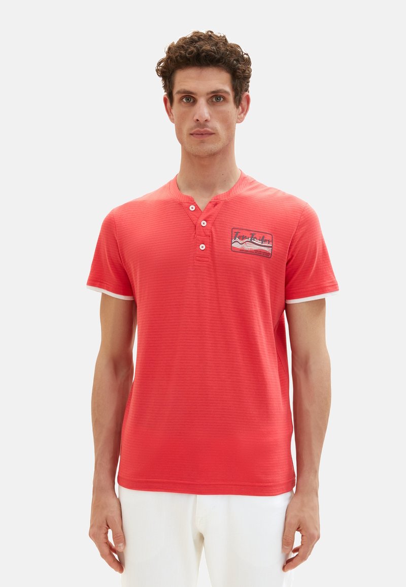Rotes gestricktes Kurzarmshirt mit Henley-Ausschnitt, zwei Knöpfen und weißen Akzenten an den Ärmeln. Mit einem aufgedruckten Logo auf der linken Brust.