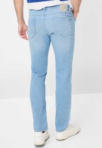 Jean en denim bleu clair avec une coupe slim, cinq poches et un patch en cuir beige marqué à l'arrière de la taille. Porté avec des baskets blanches.