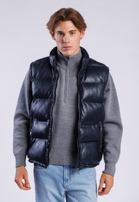 Marineblå læderpufvest med høj krave, lynlås og sidelommer, båret over en grå sweater, parret med lyseblå jeans.