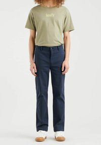 Oliwkowa zielona koszulka z logo „Levi's”, zestawiona z granatowymi spodniami, dopełniona białymi trampkami z gumową podeszwą.