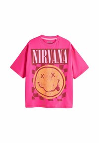 Felroze oversized T-shirt met de tekst "NIRVANA" en een grote versleten gele glimlach met kruiselings doorgestreepte ogen op een geruit achtergrond.