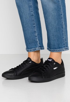 Osoba ubrana w niebieskie dżinsy i czarne trampki Puma stoi na jasnej podłodze na białym tle.