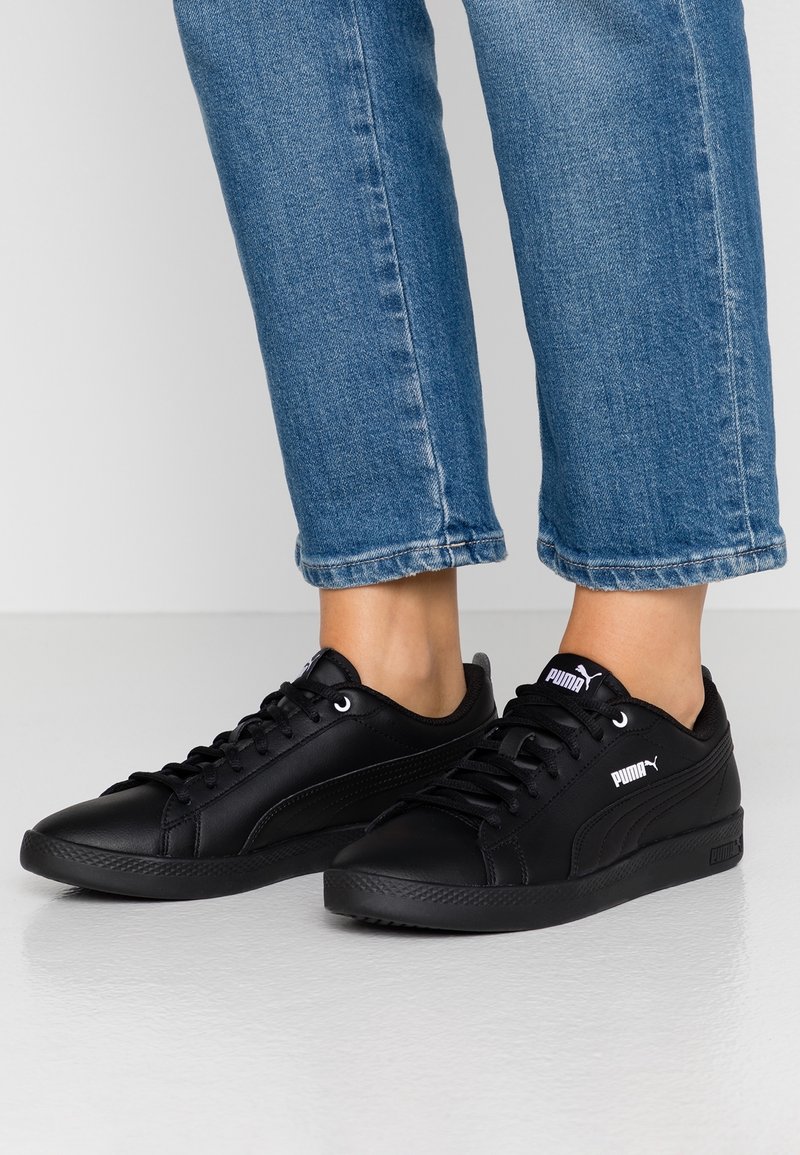 Personne portant un jean bleu et des baskets noires Puma, debout sur un sol clair avec un fond blanc.