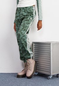 Zelené joggingové kalhoty s tie-dye vzorem a bílými pruhy, s elastickým pasem, a růžové šněrovací boty s tlustou černou podrážkou, na šedém pozadí.