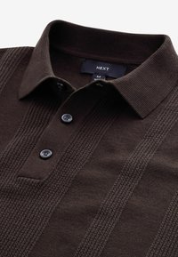 Polo marron en maille avec un motif texturé, trois boutons, et une étiquette indiquant "NEXT" ainsi que la taille "M" au col.