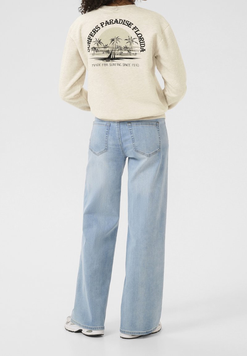 Sudadera beige con estampado gráfico negro de palmeras y un velero. Jeans de pierna ancha en azul claro. Estilo casual con texturas suaves.