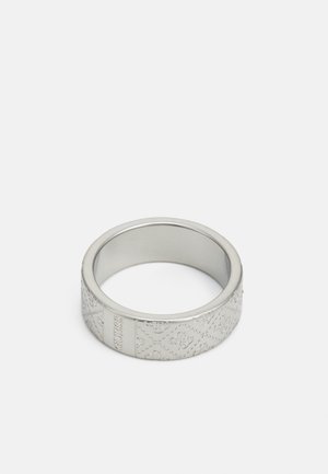 Anneau en métal ton argent gravé d'un motif de logo répétitif et du mot "GUESS" sur un fond blanc uni.
