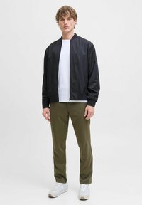 Veste bomber noire avec fermeture éclair, associée à un t-shirt blanc, un pantalon vert olive et des baskets blanches. Tissu lisse, design simple.