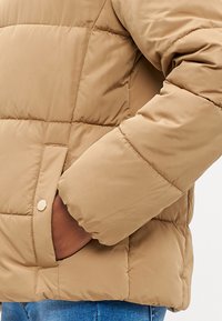 Bežna pufasta jakna s prešito teksturo, ki ima stransko žep in gladko površino. Manšeta je nekoliko nabrana za udobje.