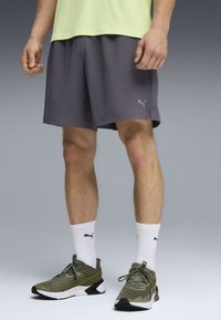 Άνδρας που φοράει γκρι σορτς Puma, λευκές κάλτσες Puma και ελιά πράσινα παπούτσια Puma, στέκεται σε ουδέτερο γκρι φόντο.