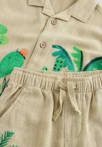Beżowa koszula zapinana na guziki i spodnie z elastycznym pasem, na przodzie koszuli nadruki zielonych i pomarańczowych kaktusów oraz liści.