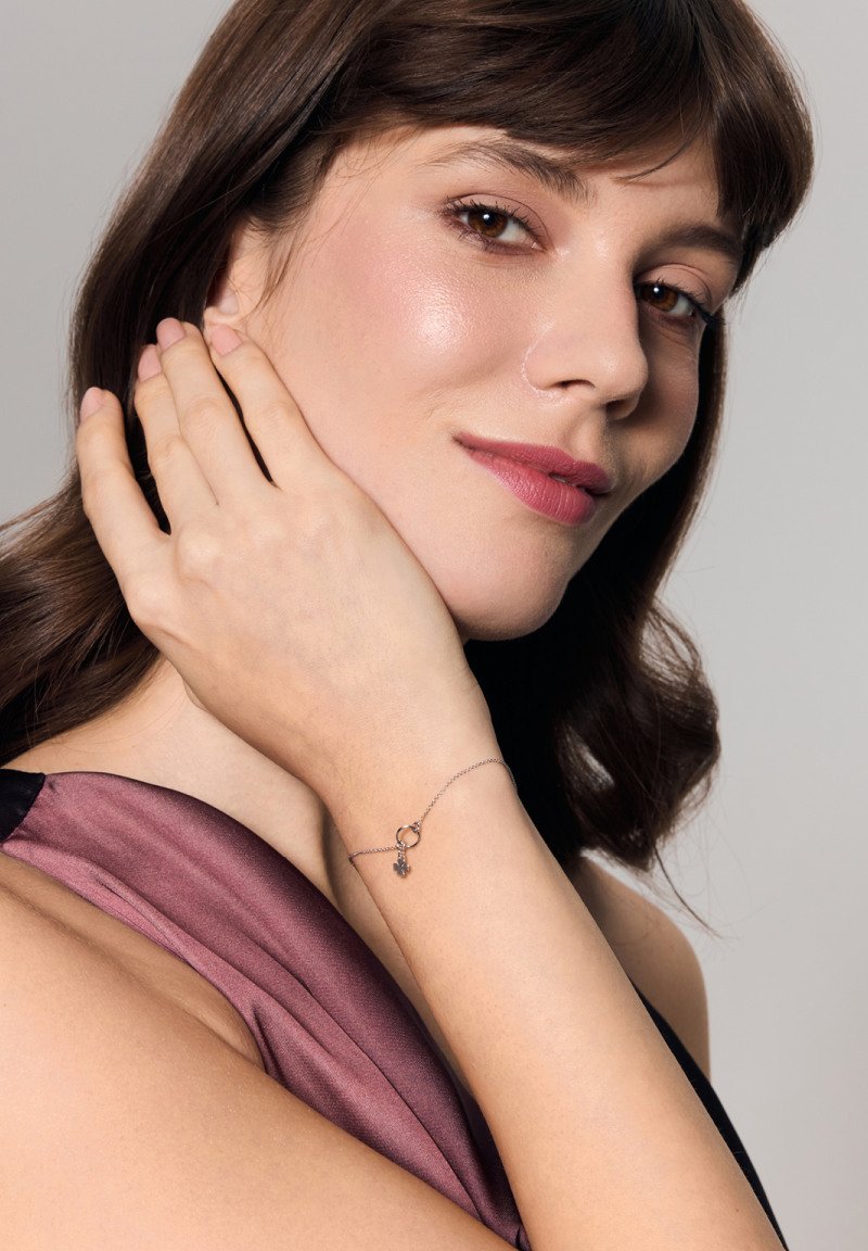 Srebrna bransoletka z okrągłym zapięciem i zawieszką, prezentowana na ręce. Tło z miękko układającego się satynowego materiału w kolorze mauve.