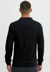 Black waffle-knit long-sleeve polo with a collared neckline, ribbed cuffs, and hem. The fabric features a textured pattern.
