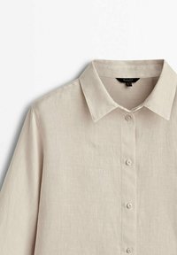 Chemise beige à manches longues avec boutons et col classique, montrée de la poitrine vers le haut sur un fond blanc.