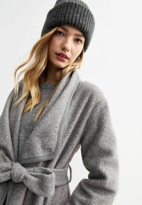 Grå ullkappa med bred krage, knytmidja och strukturerad yta. I kombination med en rippad mörkgrå mössa. Minimalistisk design och mjukt material.