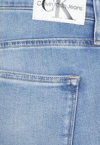 Ljusblå denimjeans med en slät textur, med en CK-logotyp på midjebandet och en ficka med kontrasterande sömnadsdetaljer.