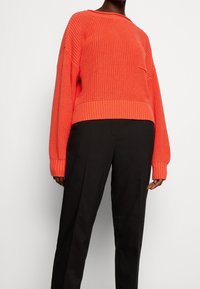 Jersey de punto naranja con mangas largas y puños acanalados, combinado con pantalones negros. El jersey presenta un diseño texturizado y un pequeño bolsillo en la parte frontal.
