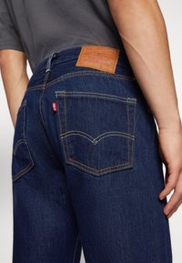 Personne portant un jean Levi's bleu foncé avec des poches arrière visibles, des coutures apparentes et une étiquette en cuir de la marque au-dessus de la poche droite.