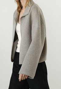 Jasnoszara, grubo teksturowana kurtka z kołnierzem i szerokimi rękawami, noszona na białej bluzce, połączona z czarnymi spodniami.