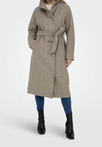 Béžový kabát z vlněné směsi s opaskem v pase, širokým límcem a dlouhými rukávy, v kombinaci s modrými úzkými džíny a černými kotníkovými botami na podpatku.