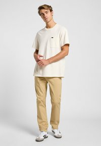 Camiseta crema de manga corta con un bolsillo en el pecho, combinada con pantalones beige claros y zapatillas blancas con franjas negras. Diseño sencillo.