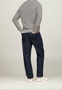 Mann in grauem geripptem Pullover, dunkelblauen Jeans und weißen Sneakers mit rot-weißen und blau-weißen Streifen, der mit Händen in den Taschen steht.