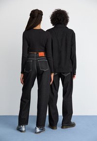 Black denim high-waisted jeans with white stitching and orange label. Model wears a fitted long-sleeve top. Casual footwear, back view.