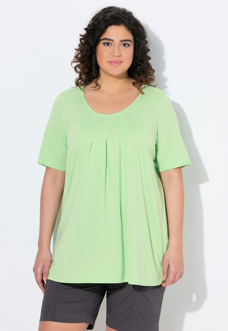 Top corto de manga corta en verde claro con escote redondo y detalle de pliegue frontal, combinado con pantalones cortos grises, exhibido sobre un fondo liso.