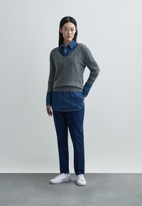 Suéter cinza de malha com tranças por cima de uma camisa de ganga, combinado com calças azul-marinho e ténis brancos. Design simples com texturas e cores em camadas.