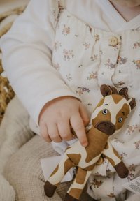 Pehmolelu norsunlepakko, jossa on ruskeita ja kermaisia laikkuja, napinmuotoiset silmät ja ruskeat kavioiset jalat, jota pitää käsi kukkakuvioisessa asussa.