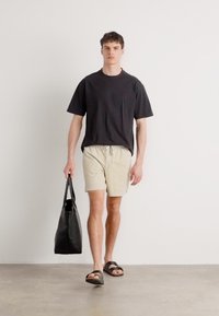 Tricou negru cu mânecă scurtă, pantaloni scurți bej și sandale negre; ținând o geantă tote neagră; fundalul este simplu și de o culoare deschisă.