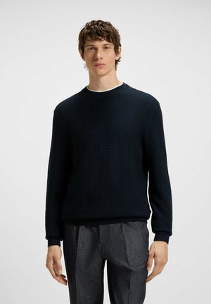 Navy textured knit sweater with a ribbed crew neckline and cuffs, paired with gray tailored trousers. Simple and modern design.