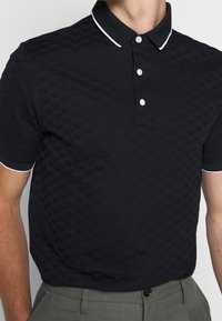 Man i svart kortärmad pikétröja med subtilt prägling och vit kantning på kragen och ärmarna, kombinerad med grå byxor.