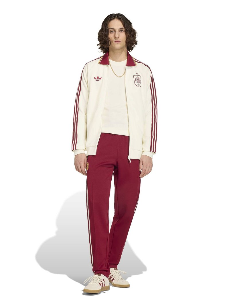 Jeune personne portant une veste de survêtement Adidas crème et bordeaux avec un pantalon assorti et des baskets coordonnées, debout devant un fond blanc.