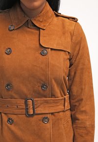 Brązowy zamszowy trencz z podwójnym zapięciem na guziki, przewiązany w pasie i z epoletami na ramionach. Charakteryzuje się gładką fakturą i wypolerowanymi guzikami.