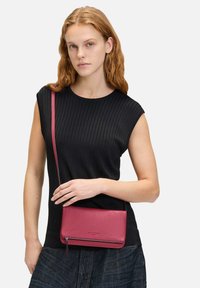 Magenta skórzana torebka crossbody o gładkiej fakturze, z zamkiem błyskawicznym i regulowanym paskiem, noszona na czarnym prążkowanym topie i dżinsach.