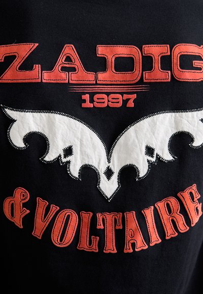 Tissu noir avec du texte rouge cousu "ZADIG 1997 & VOLTAIRE" et un design décoratif blanc en forme d'aile au centre.