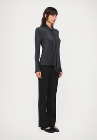 Schwarze Bluse mit einem geknoteten Detail am Kragen, kombiniert mit weit geschnittenen schwarzen Hosen und flachen Schuhen, wodurch ein monochromatischer Look entsteht.