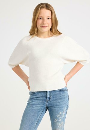 Teenage pige med lyst hår iført en hvid kortærmet sweater og blå jeans med blomsterbroderi, stående med hænderne på hofterne.