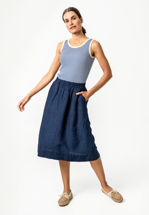 Frau trägt ein blau-weiß gestreiftes ärmelloses Oberteil, einen marineblauen Midi-Rock mit Taschen und bequeme beigen Schuhe, steht vor weißem Hintergrund.