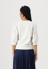 Cream pullover sweatshirt with puffed sleeves, ribbed cuffs, and hem. Worn over a long, pleated navy skirt. Smooth texture, relaxed fit.