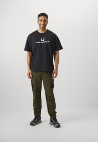 Fekete pamut póló "UNDER ARMOUR" logóval, khaki zöld melegítőnadrággal, amely fekete részletekkel és két oldalsó zsebbe rendelkezik. Szürke sportcipő.