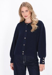 Mørkeblå strikket cardigan med rund hals, knaplukning foran og guldtonede knapper ved ærmerne. Har en afslappet pasform og struktureret stof.