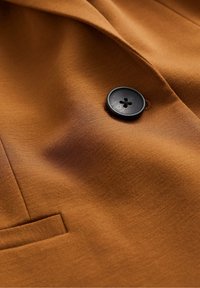 Brunt tyg med en mjuk yta, med en svart fyrhålsknapp och en liten, rundad fickdetalj.