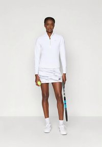 Ženska teniserka u bijeloj sportskoj odjeći drži tenisku lopticu i reketa, stojeći nasuprot jednobojnoj svijetloj pozadini.