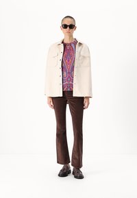 Fehér gombos dzseki, élénk paisley pulóver, barna kordbársony trapéznadrág, fekete napszemüveg, barna cipők, minimalista háttér.