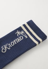 Sötétkék bordázott zokni fehér csíkokkal, kiemelkedő "KAOTIKO" felirattal és pálmafával díszítve. Az anyag lágy és lélegzőnek tűnik.