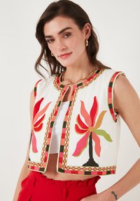 Débardeur blanc sans manches avec broderies florales en rouge, orange, vert et noir ; présente un biais à motif et un col rond.