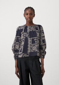 Chemisier bleu marine avec des motifs géométriques et floraux crémeux, encolure ronde et manches bouffantes, associé à un pantalon large noir.